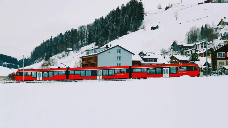 A vibrant red train traverses a snowy winter landscape against a backdrop of hills and houses.